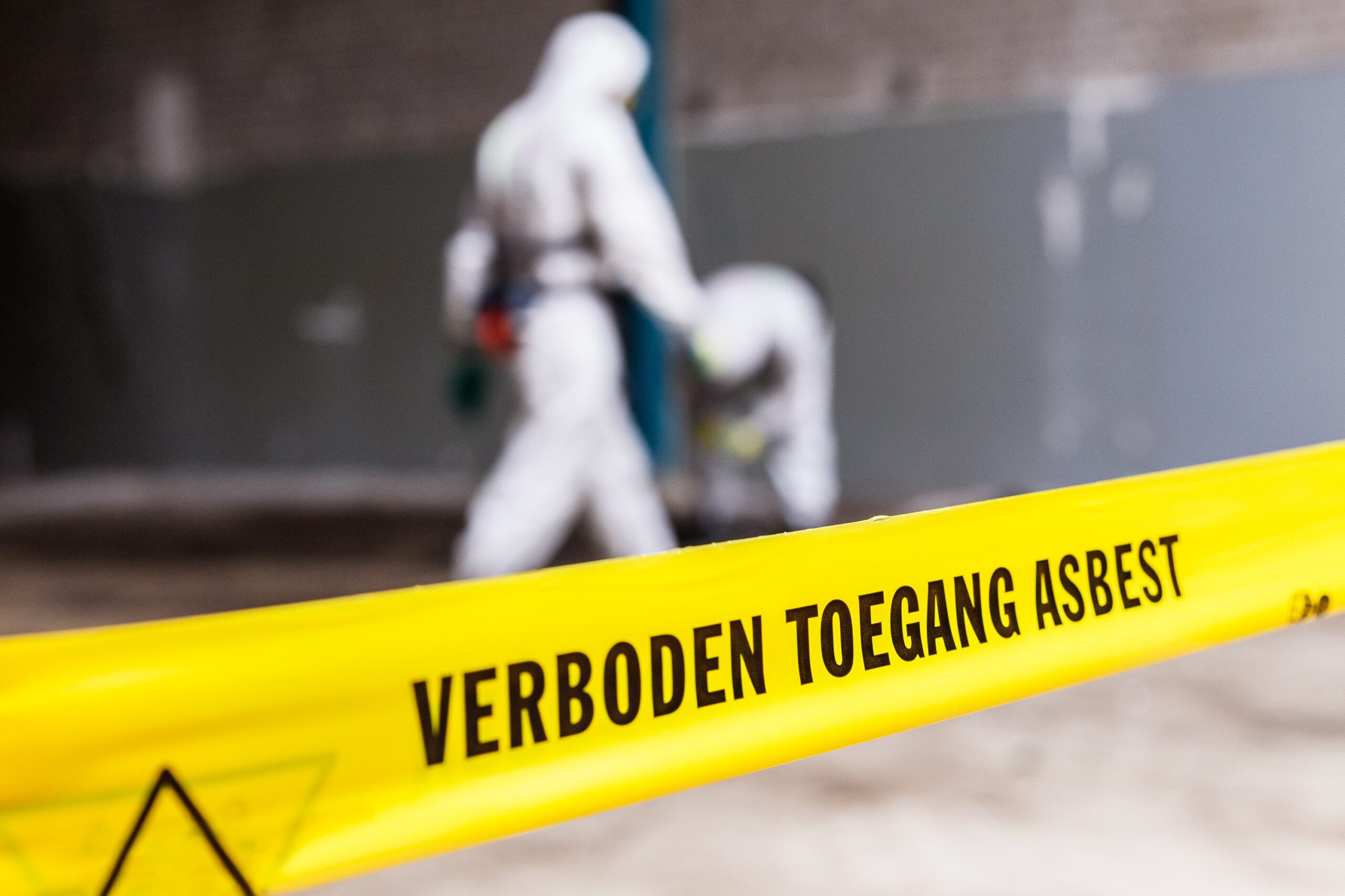 Geel alsbestlint met de tekst 'verboden toegang asbest' met op de achtergrond twee mannen in witte pakken.